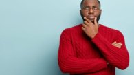 Human facial expressions concept. Contemplative dark skinned guy rubs chin as thinks, makes decision, focused away, keeps hands partly crossed, dressed in knitted jumper, models over blue wall
