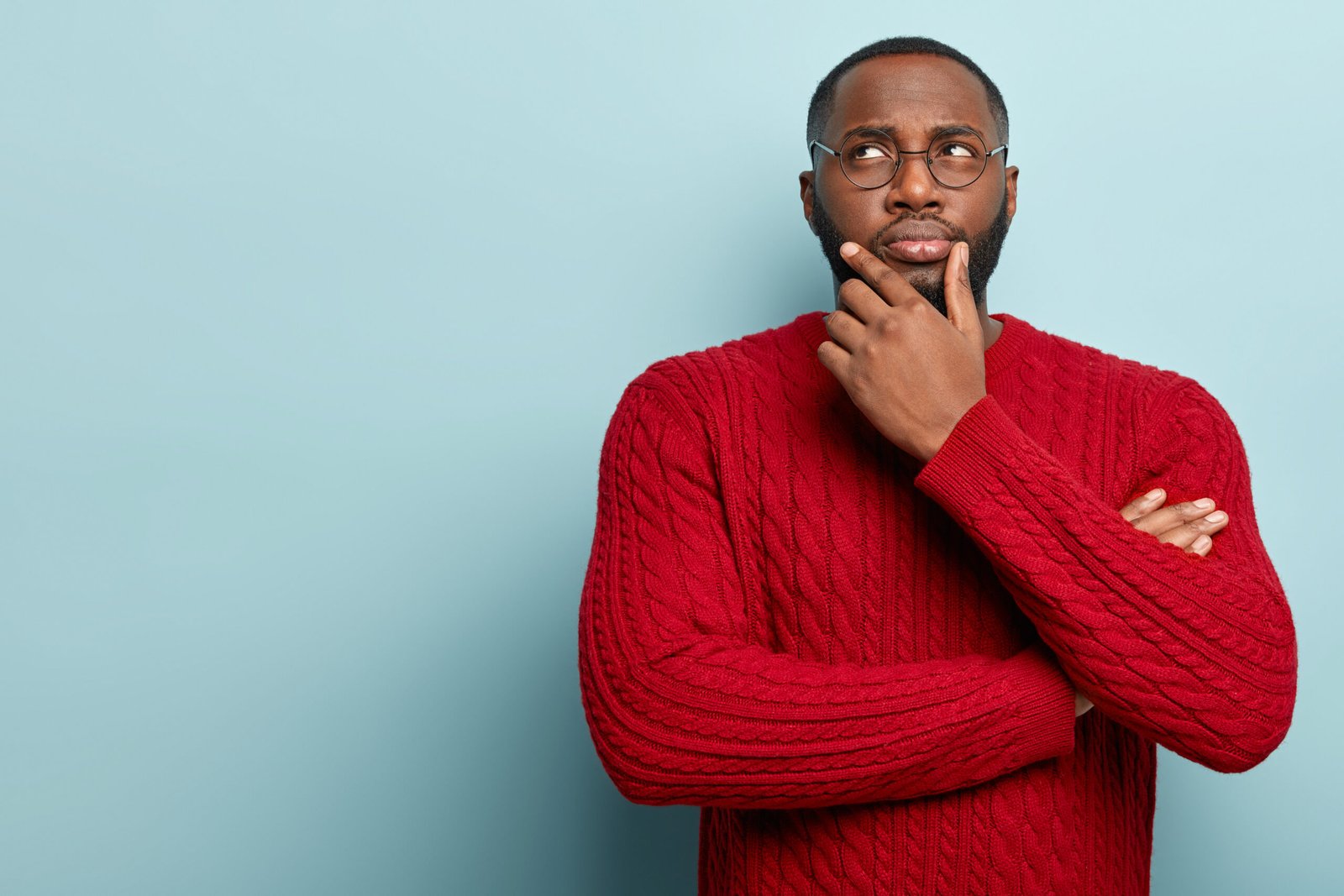 Human facial expressions concept. Contemplative dark skinned guy rubs chin as thinks, makes decision, focused away, keeps hands partly crossed, dressed in knitted jumper, models over blue wall